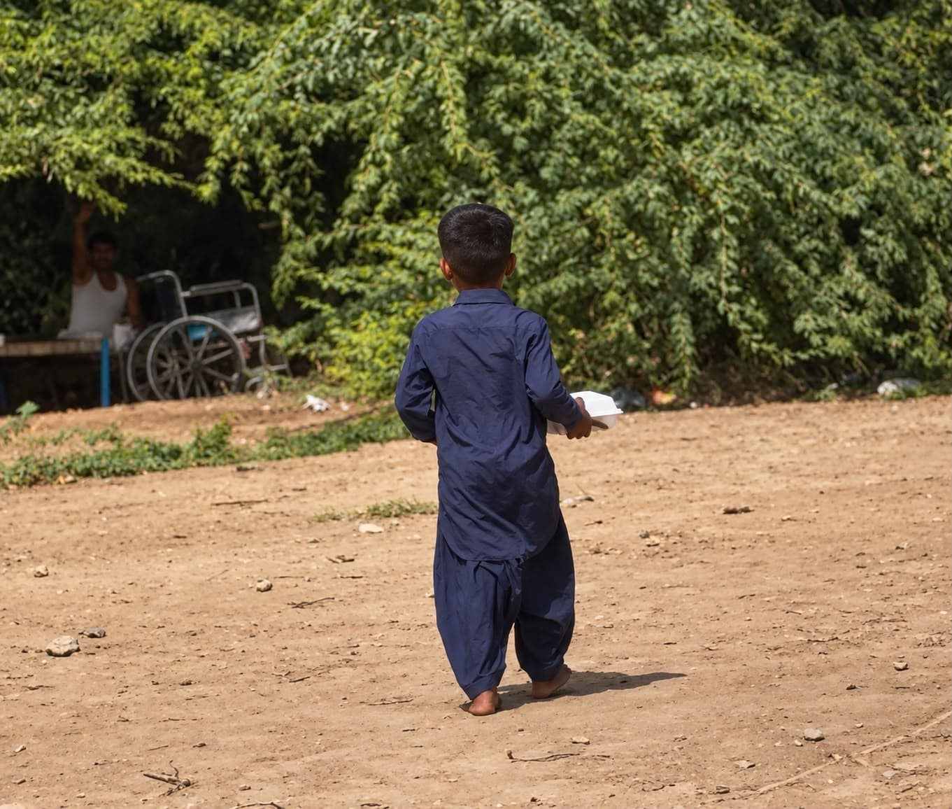 A young boy walking away along a dirt path, carrying his meal box